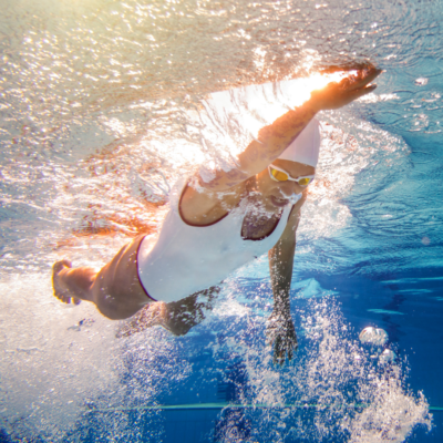 Alt: "Underwater view of a swimmer in a white cap and swimsuit reaching forward in a freestyle stroke, with sunlight filtering through the water and creating a radiant effect around the dynamic movement.