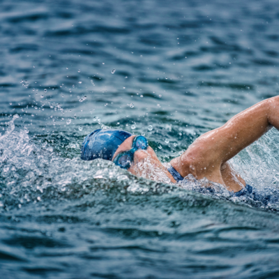 A focused triathlete in a wetsuit, goggles, and a swim cap powerfully swimming freestyle in open water, creating splashes with each stroke